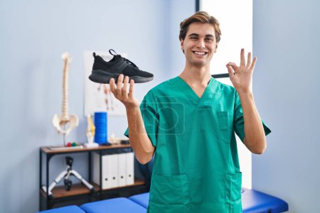Photo for Young man working at physiotherapy clinic holding shoe doing ok sign with fingers, smiling friendly gesturing excellent symbol - Royalty Free Image