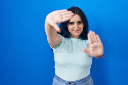 Téléchargez les photos : Jeune fille moderne avec des cheveux bleus debout sur fond bleu faisant cadre en utilisant les mains paumes et les doigts, perspective de la caméra - en image libre de droit