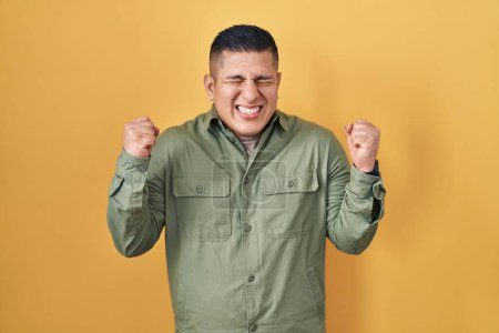 Photo for Hispanic young man standing over yellow background excited for success with arms raised and eyes closed celebrating victory smiling. winner concept. - Royalty Free Image