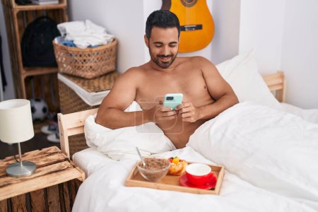 Téléchargez les photos : Young hispanic man using smartphone having breakfast sitting on bed at bedroom - en image libre de droit