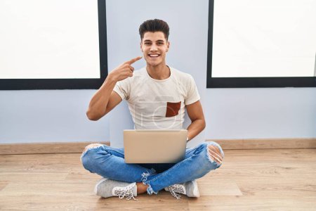 Téléchargez les photos : Jeune homme hispanique à l'aide d'un ordinateur portable à la maison souriant joyeuse montrant et pointant du doigt les dents et la bouche. concept de santé dentaire. - en image libre de droit