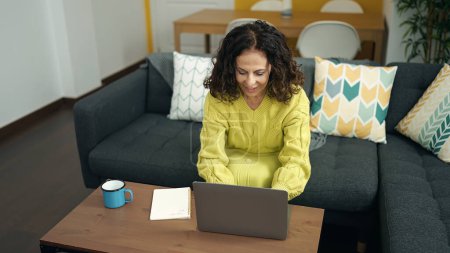 Téléchargez les photos : Moyen Age femme hispanique en utilisant un ordinateur portable assis sur le canapé à la maison - en image libre de droit