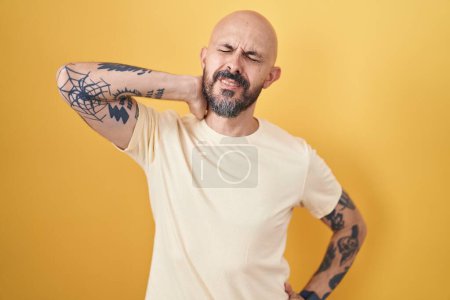 Foto de Hombre hispano con tatuajes de pie sobre fondo amarillo sufriendo de dolor de cuello lesión, tocando el cuello con la mano, dolor muscular - Imagen libre de derechos