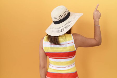 Foto de Mujer china de mediana edad con sombrero de verano sobre fondo amarillo posando hacia atrás apuntando hacia adelante con la mano del dedo - Imagen libre de derechos