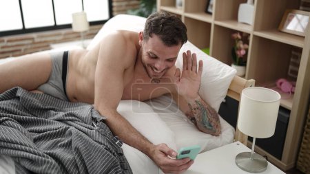 Foto de Young caucasian man doing video call with smartphone lying on the bed at bedroom - Imagen libre de derechos