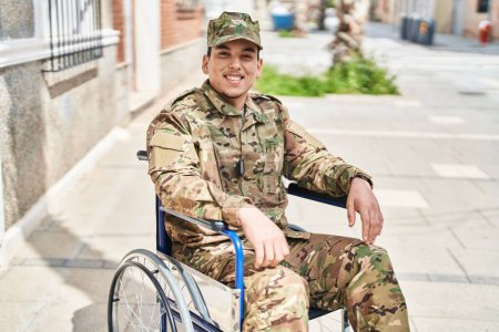 Téléchargez les photos : Jeune homme arabe portant un uniforme de camouflage de l'armée assis sur un fauteuil roulant regardant positif et heureux debout et souriant avec un sourire confiant montrant les dents - en image libre de droit
