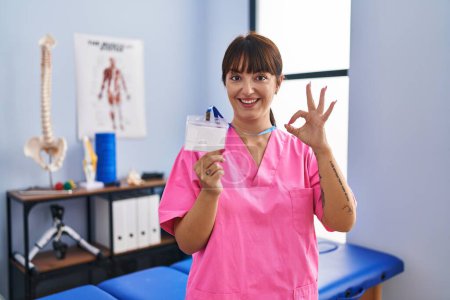 Photo for Young brunette woman working at rehabilitation clinic showing id doing ok sign with fingers, smiling friendly gesturing excellent symbol - Royalty Free Image