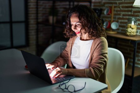 Foto de Mujer hispana de mediana edad usando laptop por la noche en casa - Imagen libre de derechos
