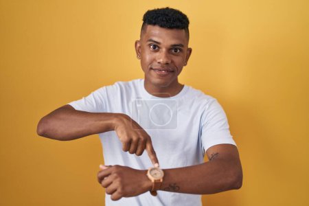 Photo for Young hispanic man standing over yellow background in hurry pointing to watch time, impatience, upset and angry for deadline delay - Royalty Free Image