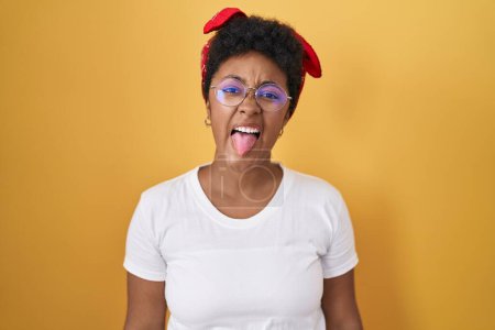 Foto de Joven mujer afroamericana de pie sobre el fondo amarillo sacando la lengua feliz con expresión divertida. concepto de emoción. - Imagen libre de derechos