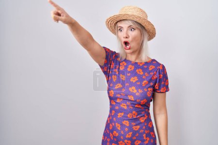 Téléchargez les photos : Jeune femme caucasienne portant des fleurs robe et chapeau d'été pointant du doigt surpris devant, bouche ouverte expression étonnée, quelque chose sur le devant - en image libre de droit