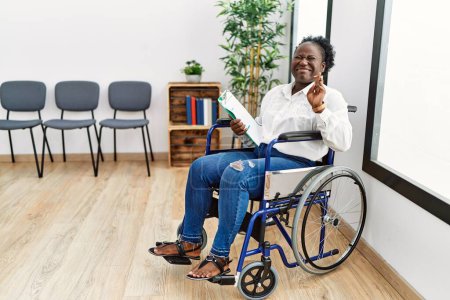 Téléchargez les photos : Jeune femme noire assise en fauteuil roulant dans la salle d'attente geste doigt croisé souriant avec espoir et les yeux fermés. chance et concept superstitieux. - en image libre de droit