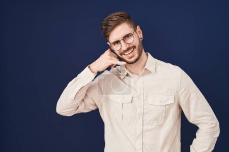 Foto de Hombre hispano con barba de pie sobre fondo azul sonriendo haciendo gesto telefónico con la mano y los dedos como hablando por teléfono. comunicar conceptos. - Imagen libre de derechos