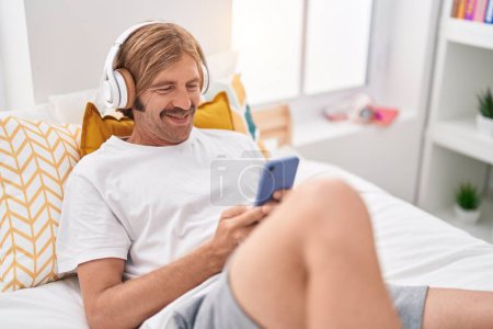 Téléchargez les photos : Jeune homme blond regarder la vidéo sur smartphone couché sur le lit dans la chambre - en image libre de droit