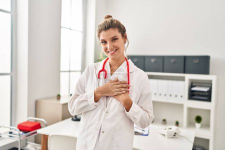 Foto de Mujer rubia joven vistiendo uniforme médico de pie con las manos en el corazón en la clínica - Imagen libre de derechos