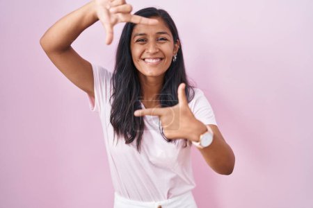 Foto de Mujer hispana joven de pie sobre fondo rosa sonriendo haciendo marco con las manos y los dedos con la cara feliz. concepto de creatividad y fotografía. - Imagen libre de derechos