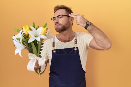 Téléchargez les photos : Homme d'âge moyen avec magasin de fleuriste barbe tenant des fleurs tirant et se suicidant pointant la main et les doigts vers la tête comme une arme à feu, geste de suicide. - en image libre de droit