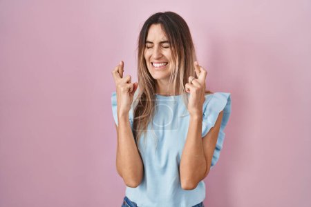 Téléchargez les photos : Jeune femme hispanique debout sur fond rose geste doigt croisé souriant avec espoir et les yeux fermés. chance et concept superstitieux. - en image libre de droit