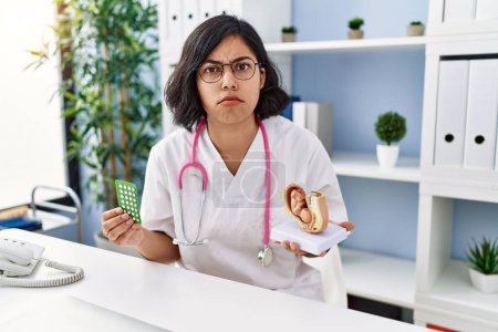 Photo for Young hispanic doctor woman holding anatomical model of uterus with fetus and birth control pills clueless and confused expression. doubt concept. - Royalty Free Image