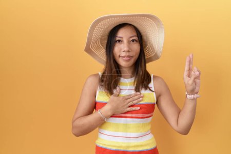 Foto de Mujer china de mediana edad con sombrero de verano sobre fondo amarillo sonriendo jurando con la mano en el pecho y los dedos hacia arriba, haciendo un juramento de lealtad promesa - Imagen libre de derechos