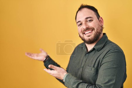 Foto de Hombre hispano de talla grande con barba de pie sobre fondo amarillo invitando a entrar sonriente natural con la mano abierta - Imagen libre de derechos