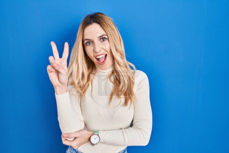 Photo for Young caucasian woman standing over blue background smiling with happy face winking at the camera doing victory sign with fingers. number two. - Royalty Free Image