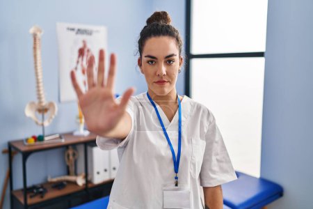 Photo for Young hispanic woman working at rehabilitation clinic doing stop sing with palm of the hand. warning expression with negative and serious gesture on the face. - Royalty Free Image