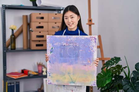 Foto de Mujer joven china sosteniendo lienzo en el estudio de arte sonriendo con una sonrisa feliz y fresca en la cara. mostrando dientes. - Imagen libre de derechos