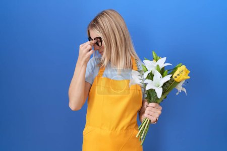 Foto de Mujer caucásica joven con delantal florista sosteniendo flores cansadas frotando la nariz y los ojos sintiendo fatiga y dolor de cabeza. concepto de estrés y frustración. - Imagen libre de derechos