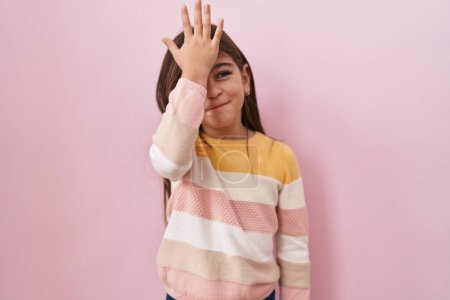 Téléchargez les photos : Petite fille hispanique portant un pull sur fond rose surpris avec la main sur la tête pour erreur, rappelez-vous erreur. oublié, mauvais concept de mémoire. - en image libre de droit