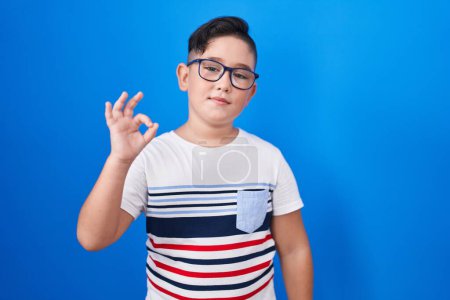 Foto de Joven niño hispano de pie sobre fondo azul sonriendo positiva haciendo signo ok con la mano y los dedos. expresión exitosa. - Imagen libre de derechos