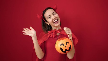 Foto de Joven hermosa mujer hispana con traje de diablo sosteniendo cesta de calabaza de Halloween sobre fondo rojo aislado - Imagen libre de derechos