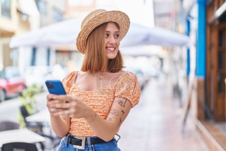Foto de Joven mujer pelirroja turista usando sombrero de verano usando smartphone en la calle - Imagen libre de derechos