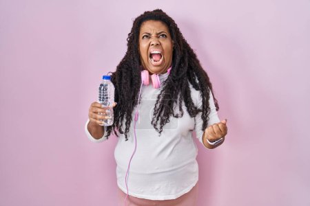 Photo for Plus size hispanic woman wearing sportswear and headphones angry and mad screaming frustrated and furious, shouting with anger. rage and aggressive concept. - Royalty Free Image