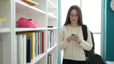 Téléchargez les photos : Jeune belle étudiante hispanique utilisant un smartphone debout à la bibliothèque de l'université - en image libre de droit