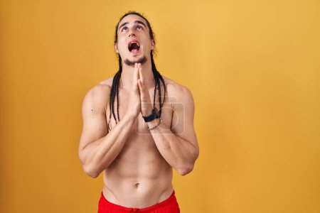 Téléchargez les photos : Homme hispanique avec les cheveux longs torse nu sur fond jaune mendier et prier avec les mains ainsi que l'expression de l'espoir sur le visage très émotionnel et inquiet. mendier. - en image libre de droit