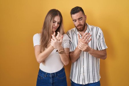 Téléchargez les photos : Jeune couple debout sur fond jaune souffrant de douleur aux mains et aux doigts, inflammation de l'arthrite - en image libre de droit
