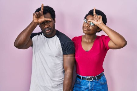 Foto de Joven pareja afroamericana de pie sobre fondo rosa burlándose de la gente con los dedos en la frente haciendo gesto perdedor burlándose e insultando. - Imagen libre de derechos