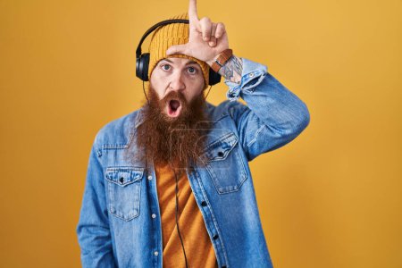 Téléchargez les photos : Homme caucasien à longue barbe écoutant de la musique à l'aide d'écouteurs se moquant des gens avec les doigts sur le front faisant geste de perdant moquerie et insultant. - en image libre de droit