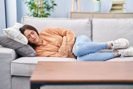 Photo for Young beautiful hispanic woman suffering for menstrual pain lying on sofa at home - Royalty Free Image