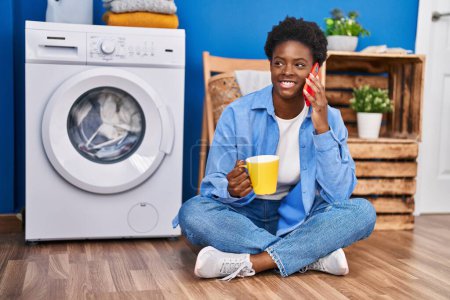 Téléchargez les photos : Femme afro-américaine parlant sur le smartphone et buvant du café attendant la machine à laver à la buanderie - en image libre de droit