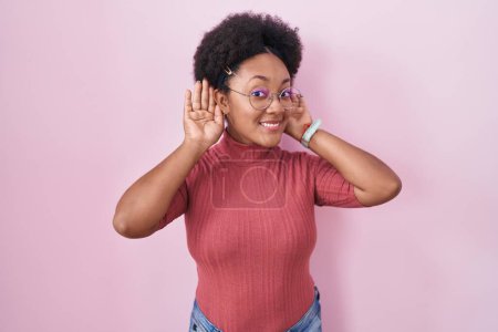 Foto de Hermosa mujer africana con el pelo rizado de pie sobre el fondo rosa tratando de escuchar ambas manos en el gesto de la oreja, curioso por chismes. problema auditivo, sordo - Imagen libre de derechos
