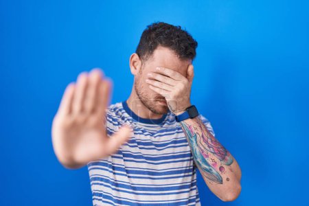 Photo for Young hispanic man standing over blue background covering eyes with hands and doing stop gesture with sad and fear expression. embarrassed and negative concept. - Royalty Free Image