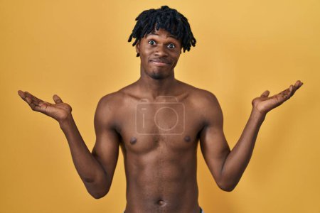 Téléchargez les photos : Jeune homme africain avec dreadlocks debout sans chemise expression désorientée et confus avec les bras et les mains levées. concept de doute. - en image libre de droit