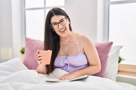 Foto de Young hispanic woman drinking cup of coffee reading book at bedroom - Imagen libre de derechos