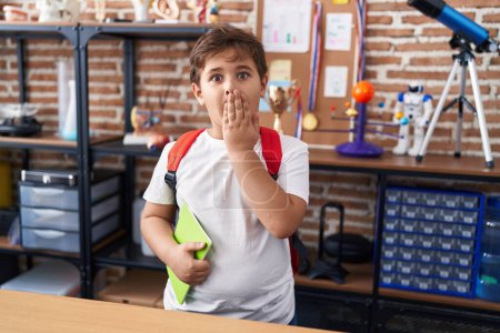 Foto de Pequeño niño hispano con mochila de estudiante y libro en la clase de la escuela cubriendo la boca con la mano, sorprendido y temeroso de equivocarse. expresión sorprendida - Imagen libre de derechos