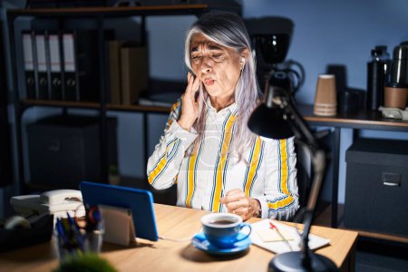 Photo for Middle age woman with grey hair working at the office at night touching mouth with hand with painful expression because of toothache or dental illness on teeth. dentist - Royalty Free Image