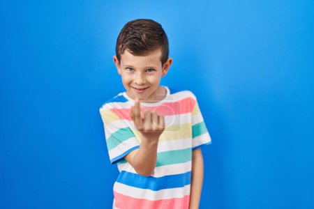Foto de Joven chico caucásico de pie sobre fondo azul haciendo señas ven aquí gesto con la mano invitando a la bienvenida feliz y sonriente - Imagen libre de derechos