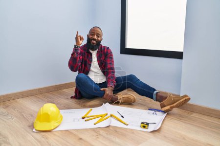 Foto de Hombre afroamericano sentado en el suelo en una casa nueva mirando los planos apuntando con el dedo hacia arriba con una idea exitosa. exitado y feliz. número uno. - Imagen libre de derechos