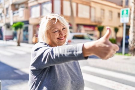 Téléchargez les photos : Moyen âge blonde femme souriante confiante faire ok signe avec le pouce vers le haut à la rue - en image libre de droit